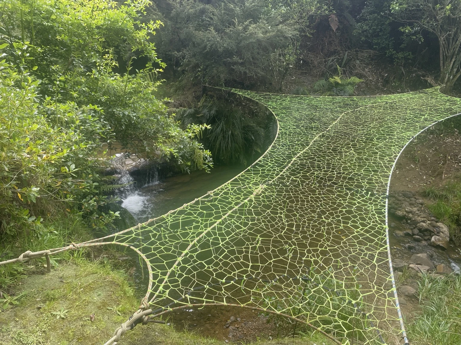 A lime-green handwoven tree net spanning a stream beside a waterfall